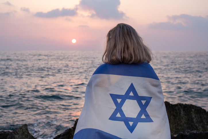 Woman looking out to sea at sunset with a flag of the State of Israel draped over her shoulders