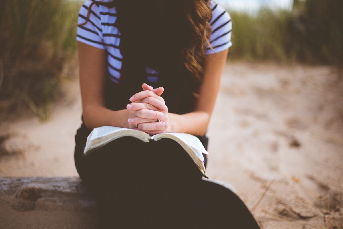 Woman seated with her clasped hands, fingers laced, resting on an open book on her lap.