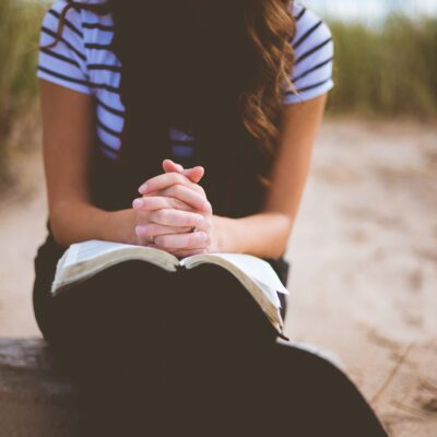 Woman seated with her clasped hands, fingers laced, resting on an open book on her lap.