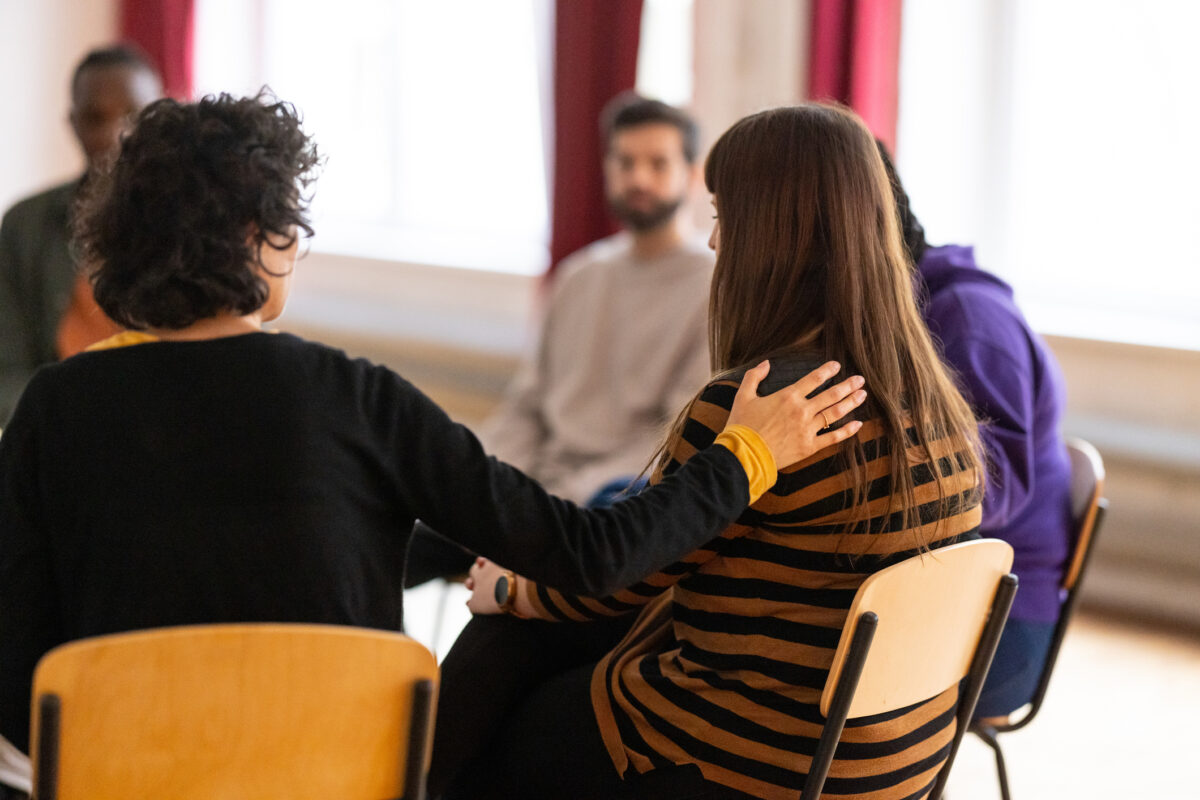 Supportive people consoling a distraught woman who is talking about her issues during group therapy meeting. Multi-ethnic group of adults attending a group therapy session.