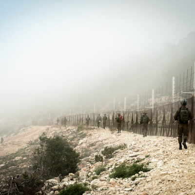 On a mountainside, soldiers walk along a fence, the mountain and sky obscured by white-gray fog.